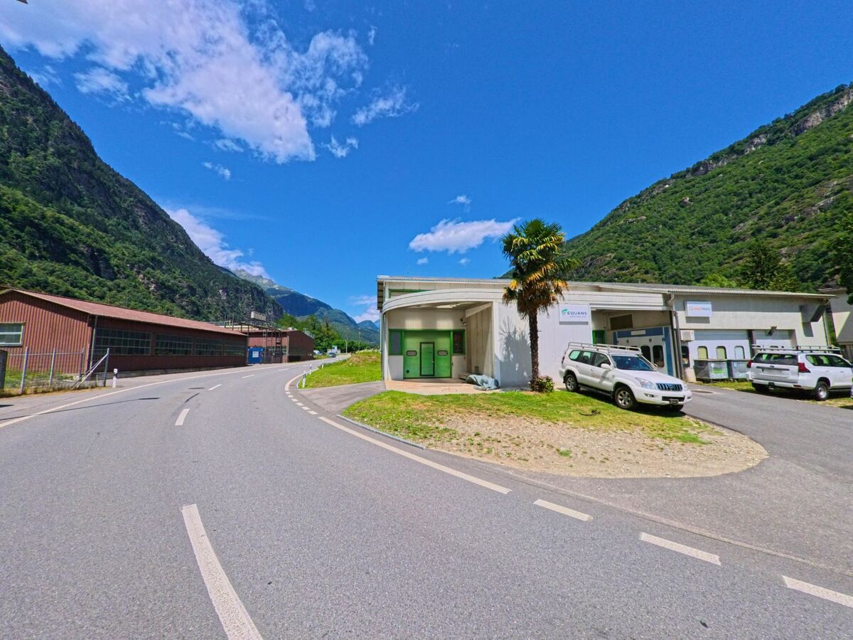 Ein helles Gebaeude mit roten Fensterlaeden steht in Bodio (6743, Kanton Tessin) oberhalb einer Strasse, neben hohen Baeumen und unter einem klaren blauen Himmel.