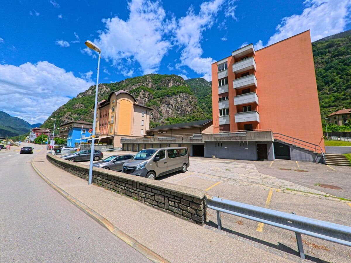 Mehrgeschossige Wohnhaeuser mit farbigen Fassaden stehen in Bodio (6743, Kanton Tessin) entlang einer breiten Strasse mit parkenden Autos und Blick auf die felsigen Berghaenge.