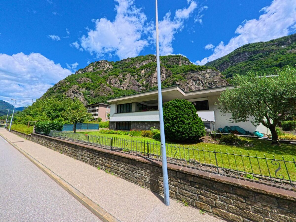 Ein modernes Wohnhaus mit flachem Dach und gepflegtem Garten steht in Bodio (6743, Kanton Tessin) vor einer steilen Felswand und unter blauem Himmel.