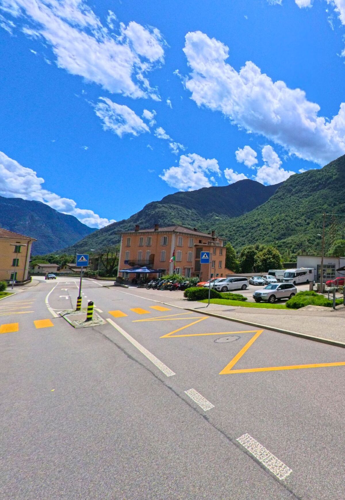 Eine Strassenkreuzung in Bodio (6743, Kanton Tessin) mit Fussgaengerstreifen und Hinweisschildern fuehrt an Wohnhaeusern vorbei, im Hintergrund ragen bewaldete Berge in den blauen Himmel.