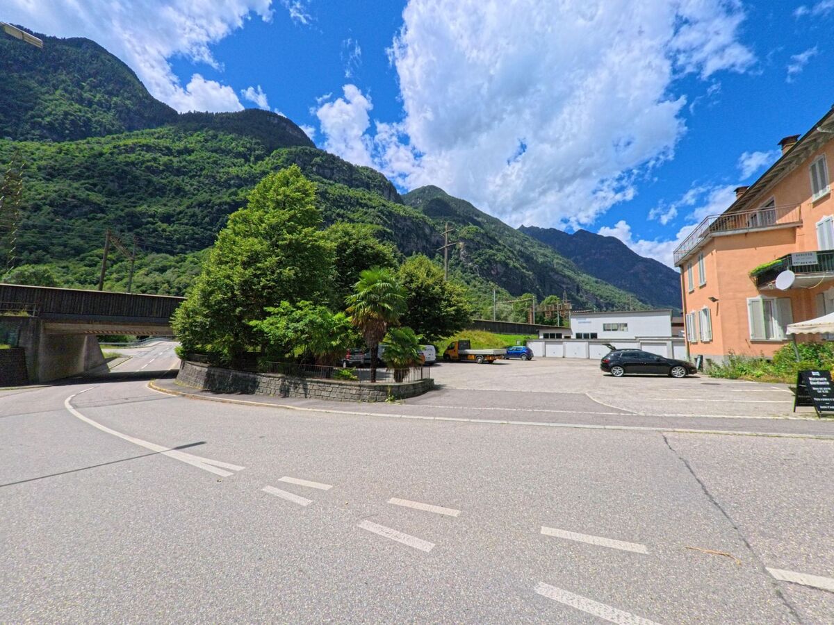 Eine Strassenkurve in Bodio (6743, Kanton Tessin) fuehrt unter einer Eisenbahnbruecke hindurch, umgeben von gruener Vegetation, Bergen und einem kleinen Parkplatz.