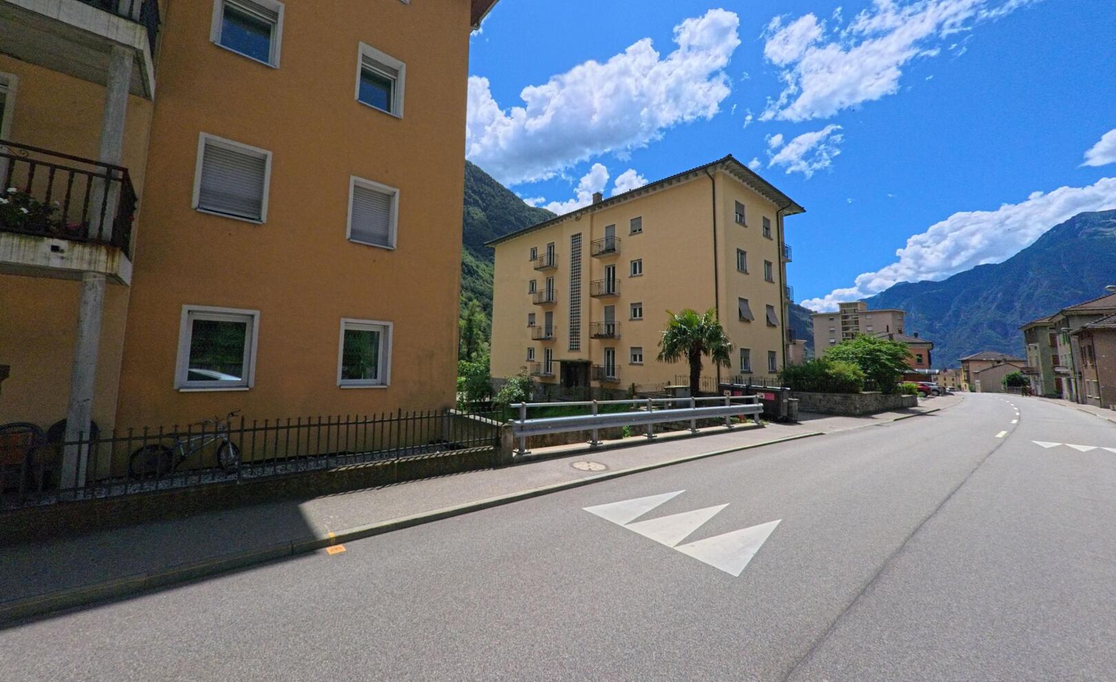 In Bodio (6743, Kanton Tessin) verlaufen Wohnhaeuser mit gelben Fassaden entlang einer ruhigen Strasse, im Hintergrund sind die Berge unter blauem Himmel sichtbar.