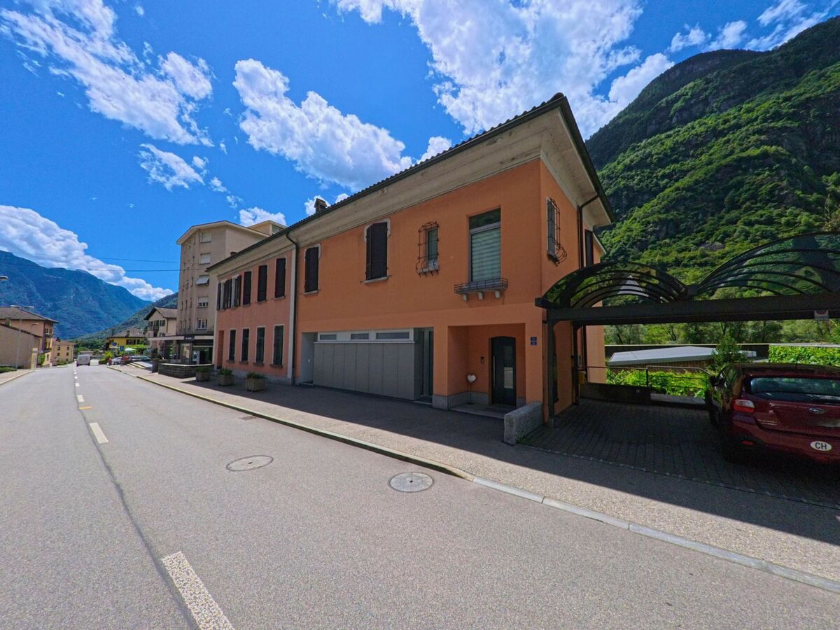 In Bodio (6743, Kanton Tessin) steht ein orangefarbenes Wohnhaus mit Garagen und Ueberdachung an einer sonnigen Strasse, umgeben von gruenen Berghaengen.