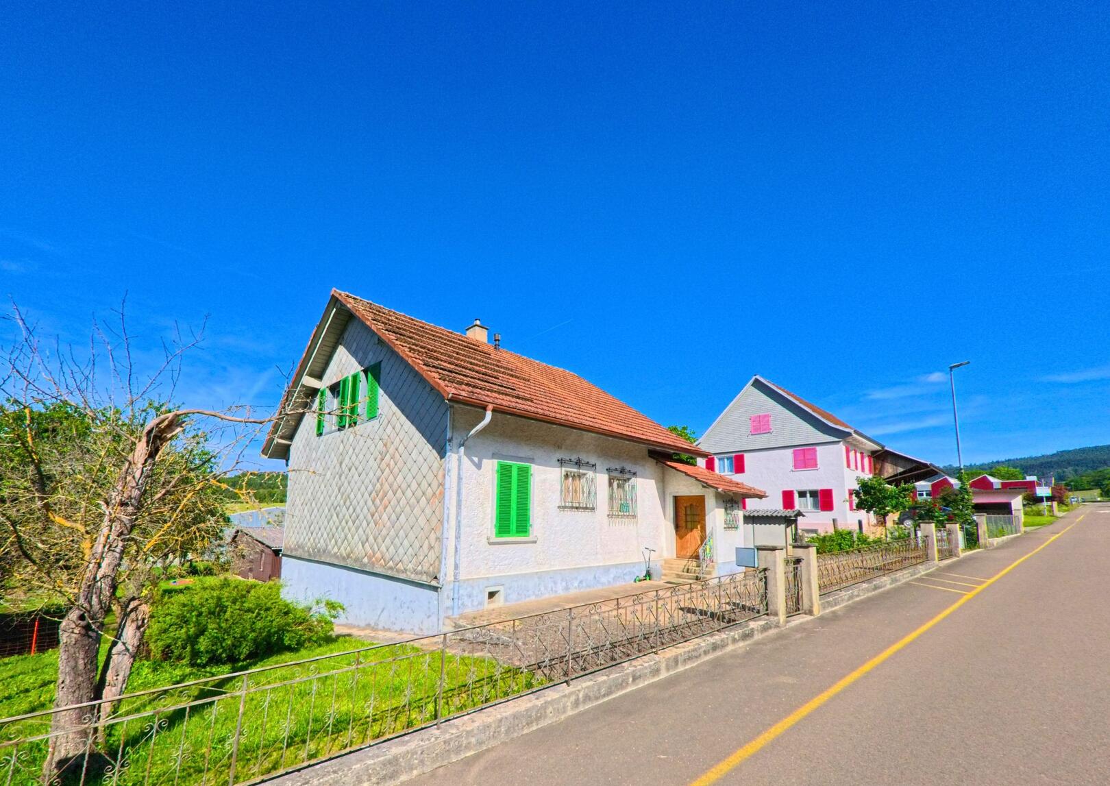 In Boécourt (2856, Kanton Jura) stehen bunte Häuser mit grünen und rosa Fensterläden entlang einer ruhigen Strasse, die von Bäumen gesäumt ist.