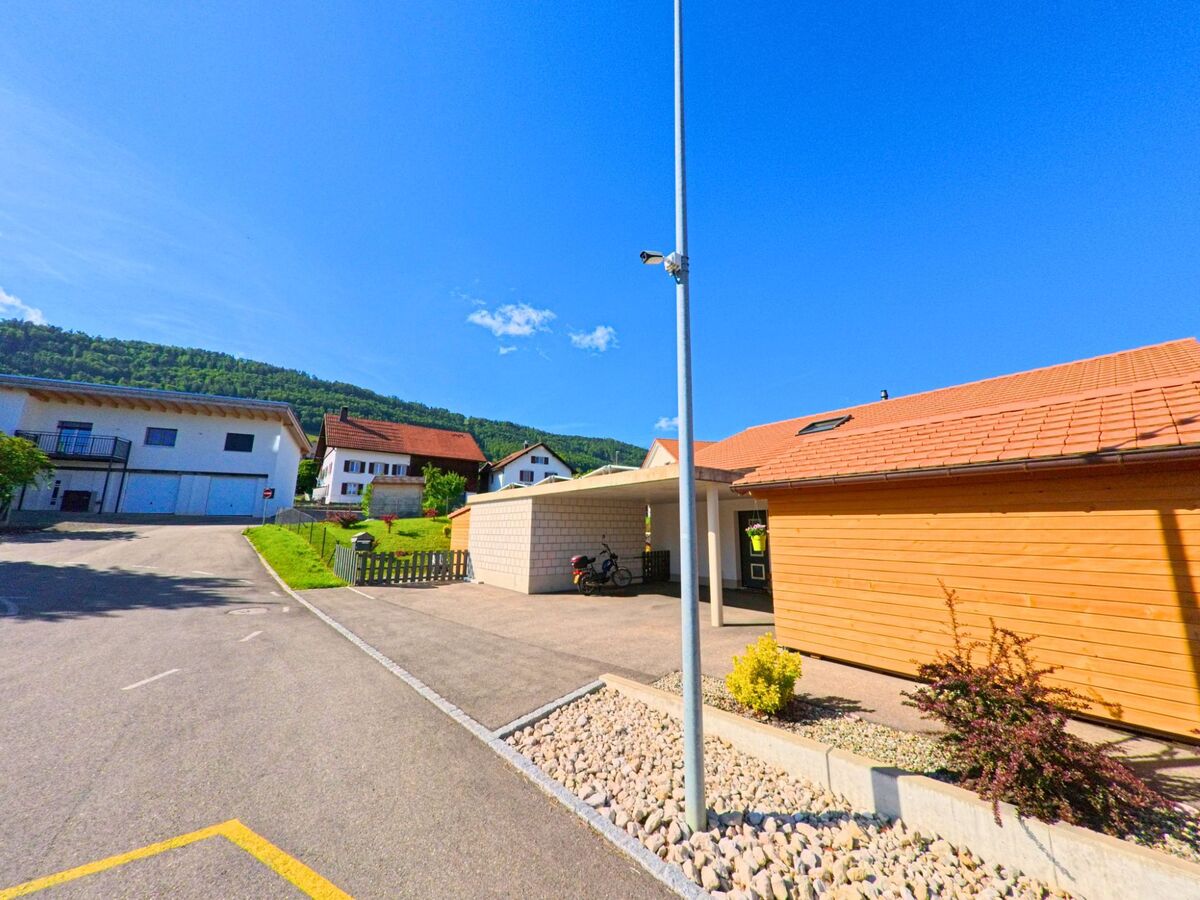 In Boécourt (2856, Kanton Jura) stehen moderne Gebäude entlang einer gut ausgebauten Straße, mit grünen Hügeln im Hintergrund und einer Straßenlaterne.