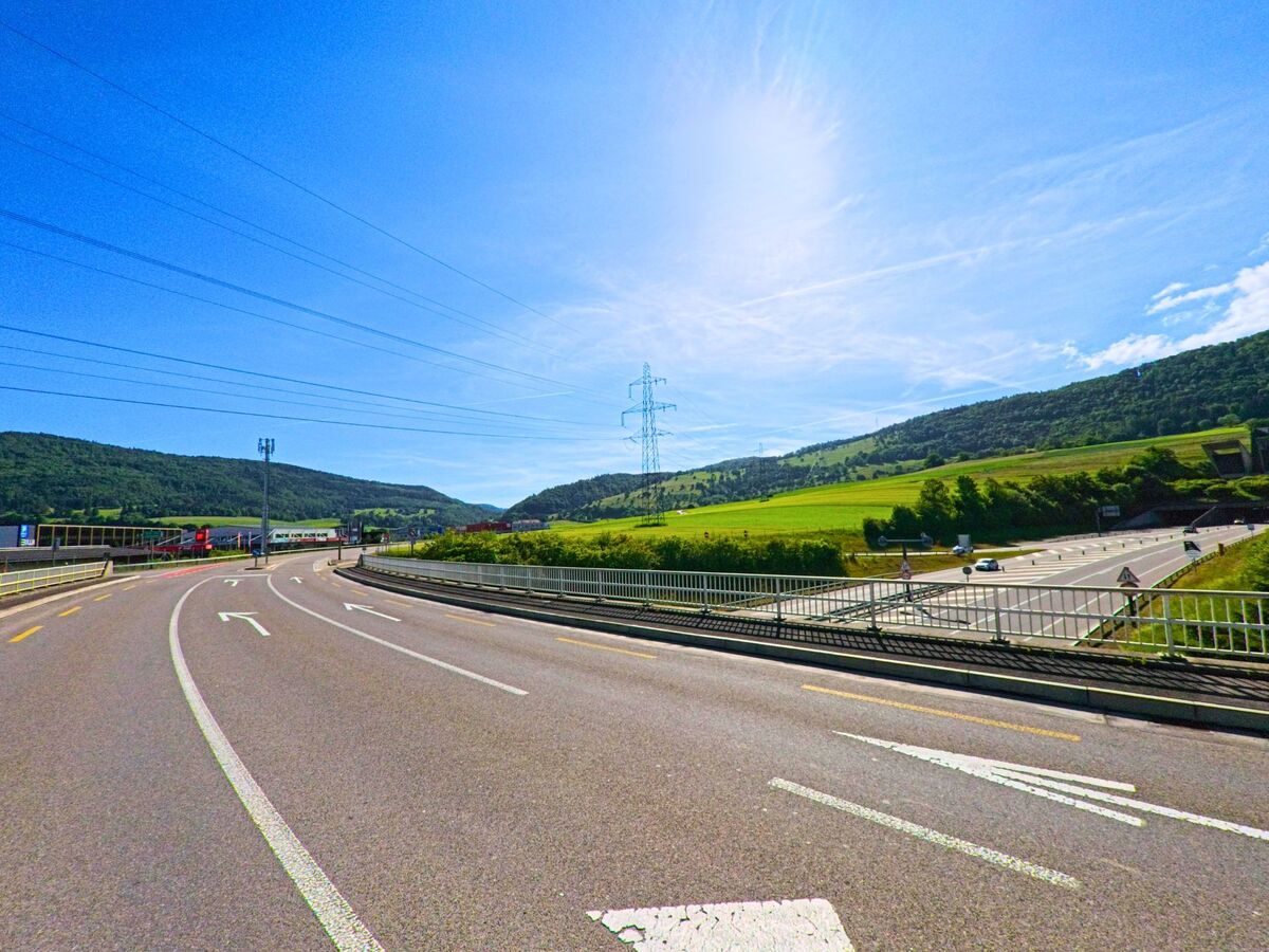 In Boécourt (2856, Kanton Jura) führt eine breite Straße durch grüne Hügel und Felder, mit Strommasten und einem klaren Himmel im Hintergrund.