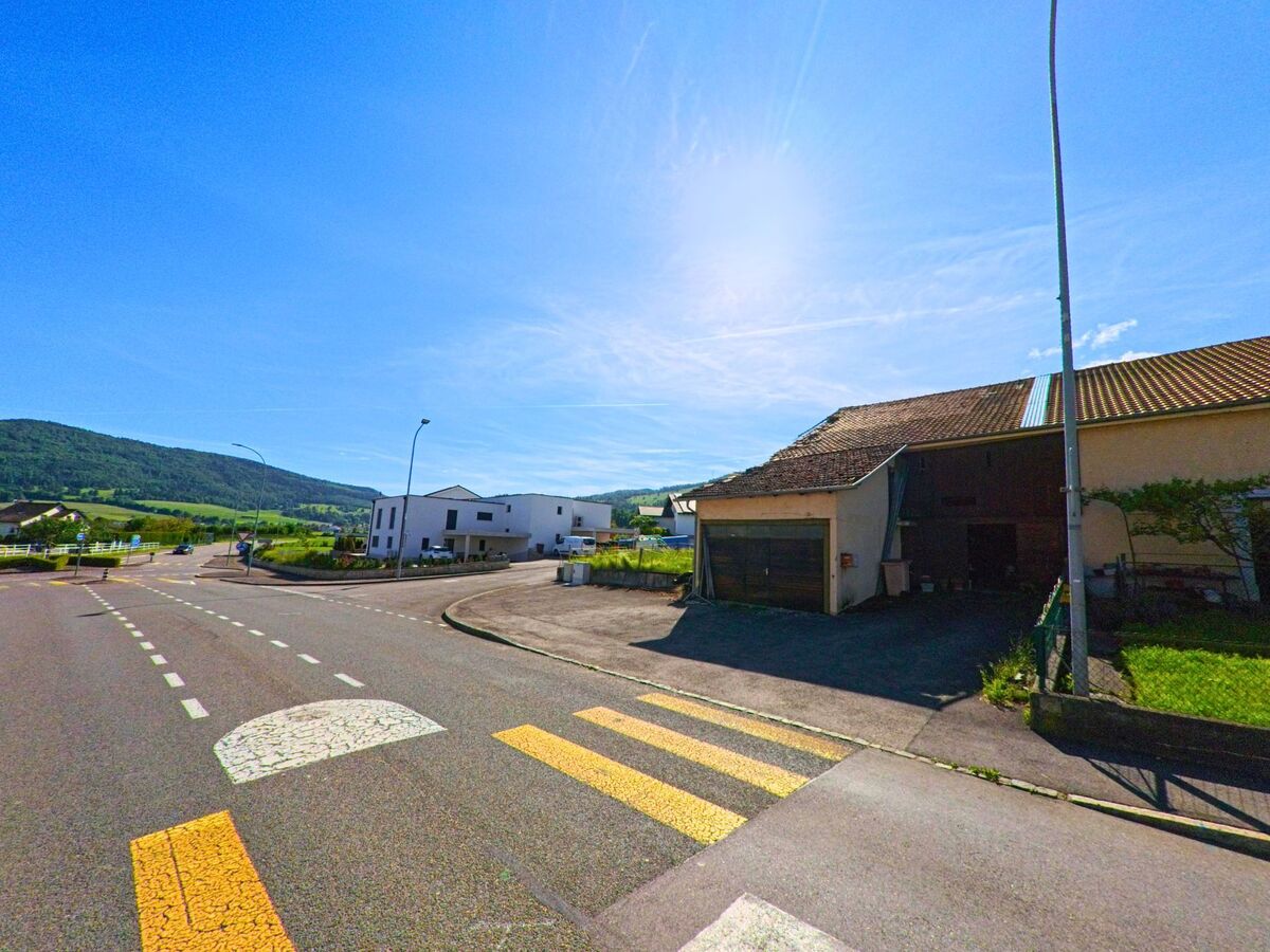 In Boécourt (2856, Kanton Jura) führt eine Straße an modernen Gebäuden und einem alten Haus entlang, mit den grünen Hügeln im Hintergrund und einer Straßenlaterne.