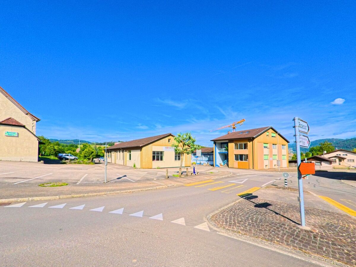 In Boécourt (2856, Kanton Jura) führt eine Straße an modernen Gebäuden und einem Parkplatz vorbei, mit einem Straßenschild und grünen Hügeln im Hintergrund.