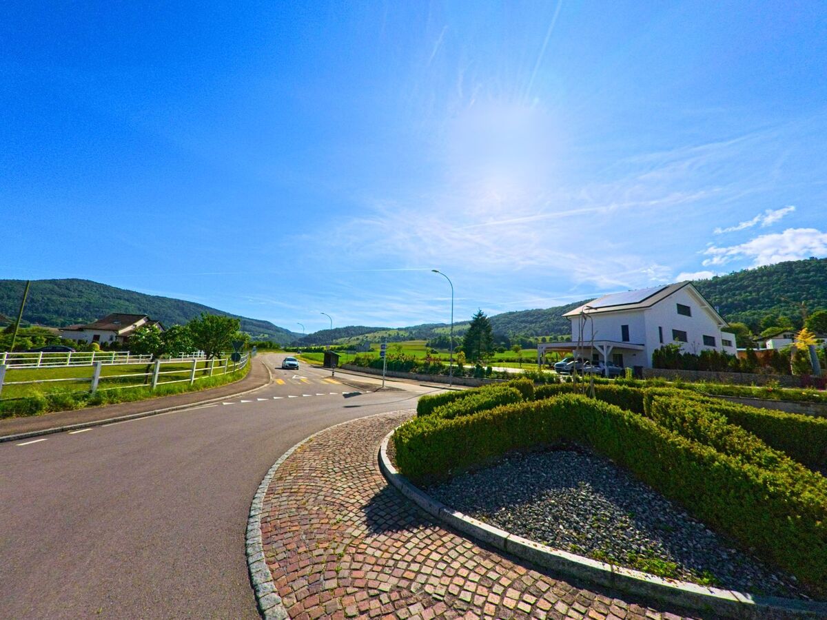 In Boécourt (2856, Kanton Jura) führt eine Straße an gepflegten Häusern und einem Garten vorbei, mit grünen Hügeln und einem klaren blauen Himmel im Hintergrund.