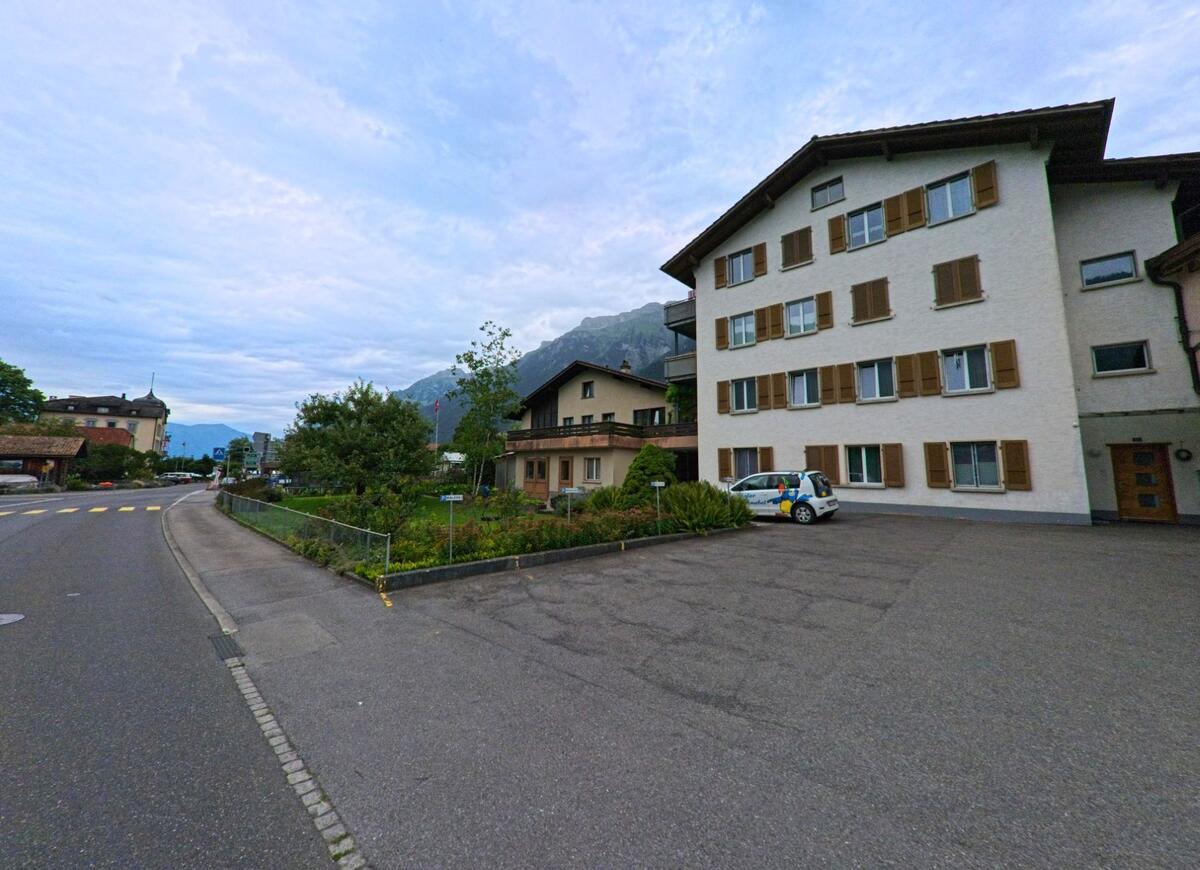 In Bönigen (3806, Kanton Bern) steht ein mehrstöckiges Wohnhaus mit Fensterläden und Parkplatz an einer Hauptstrasse mit Blick auf die umliegenden Berge.