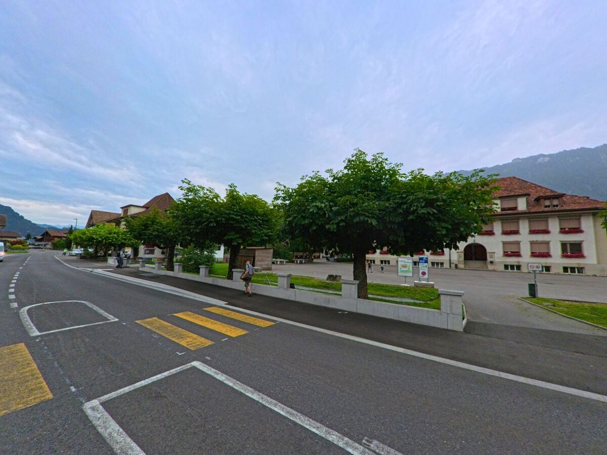 In Bönigen (3806, Kanton Bern) führt eine Hauptstrasse mit Zebrastreifen am Schulhaus vorbei, dessen Fassade mit roten Fensterläden und Blumenschmuck versehen ist.