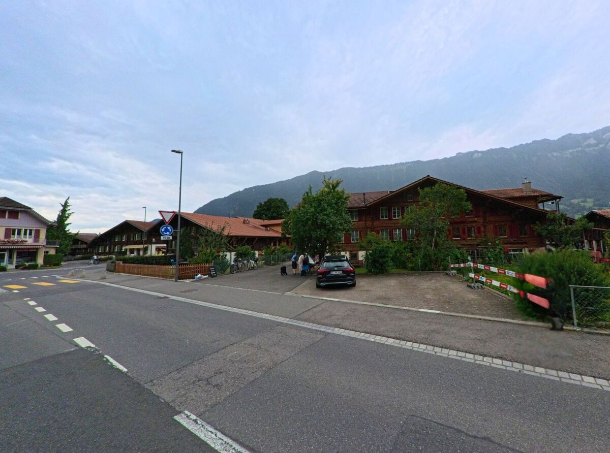 In Bönigen (3806, Kanton Bern) stehen traditionelle Holzhäuser an einem Strassenknoten mit Vorfahrtregelung, während sich einige Personen auf einem Parkplatz unterhalten.