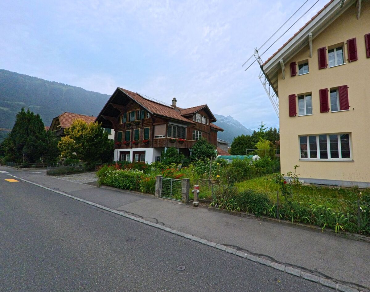 In einer Wohnstrasse von Bönigen (3806, Kanton Bern) steht ein traditionelles Berner Haus mit gepflegtem Vorgarten und roten Fensterblumen, flankiert von weiteren Wohngebäuden.