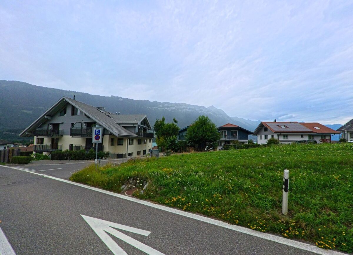 Am Ortsrand von Bönigen (3806, Kanton Bern) stehen moderne Wohnhäuser vor einer grünen Wiese mit Blick auf die umliegenden Berghänge.