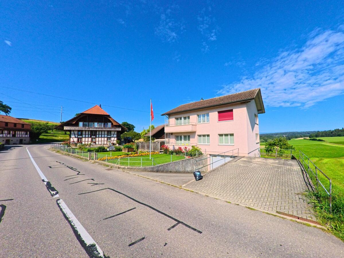 In Bösingen (3178, Kanton Freiburg) stehen ein traditionelles Fachwerkhaus und ein modernes Wohnhaus nebeneinander an einer leicht ansteigenden Strasse mit Blick auf die umliegenden Felder.