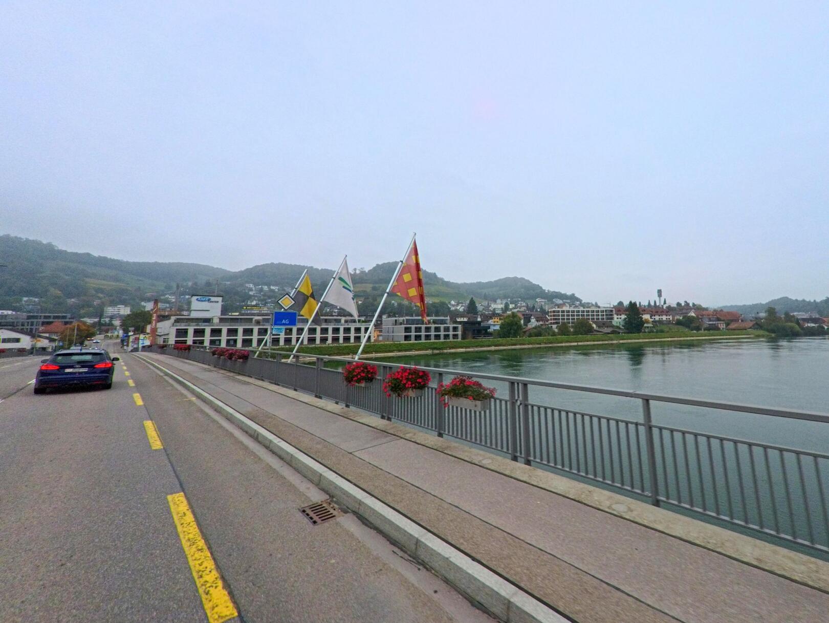 Von der Brücke über die Aare bietet sich ein Blick Richtung Döttingen, unweit von Böttstein (5312, Kanton Aargau), mit den Fahnen am Geländer und sanften Hügeln im Hintergrund.