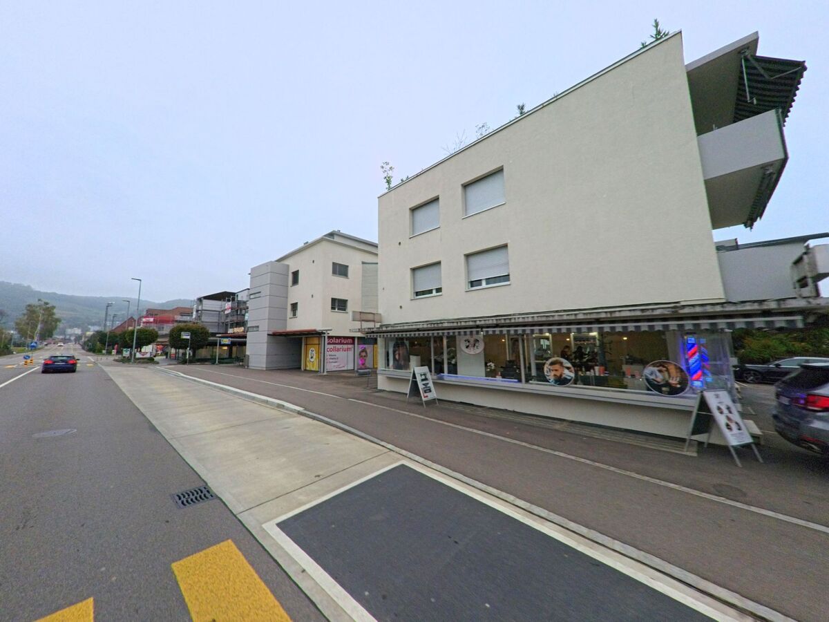 Entlang der Hauptstrasse in Böttstein (5314, Kanton Aargau) stehen moderne Gebäude mit Geschäften im Erdgeschoss, darunter ein Friseursalon und ein Solarium, vor dem Hintergrund bewaldeter Hügel.