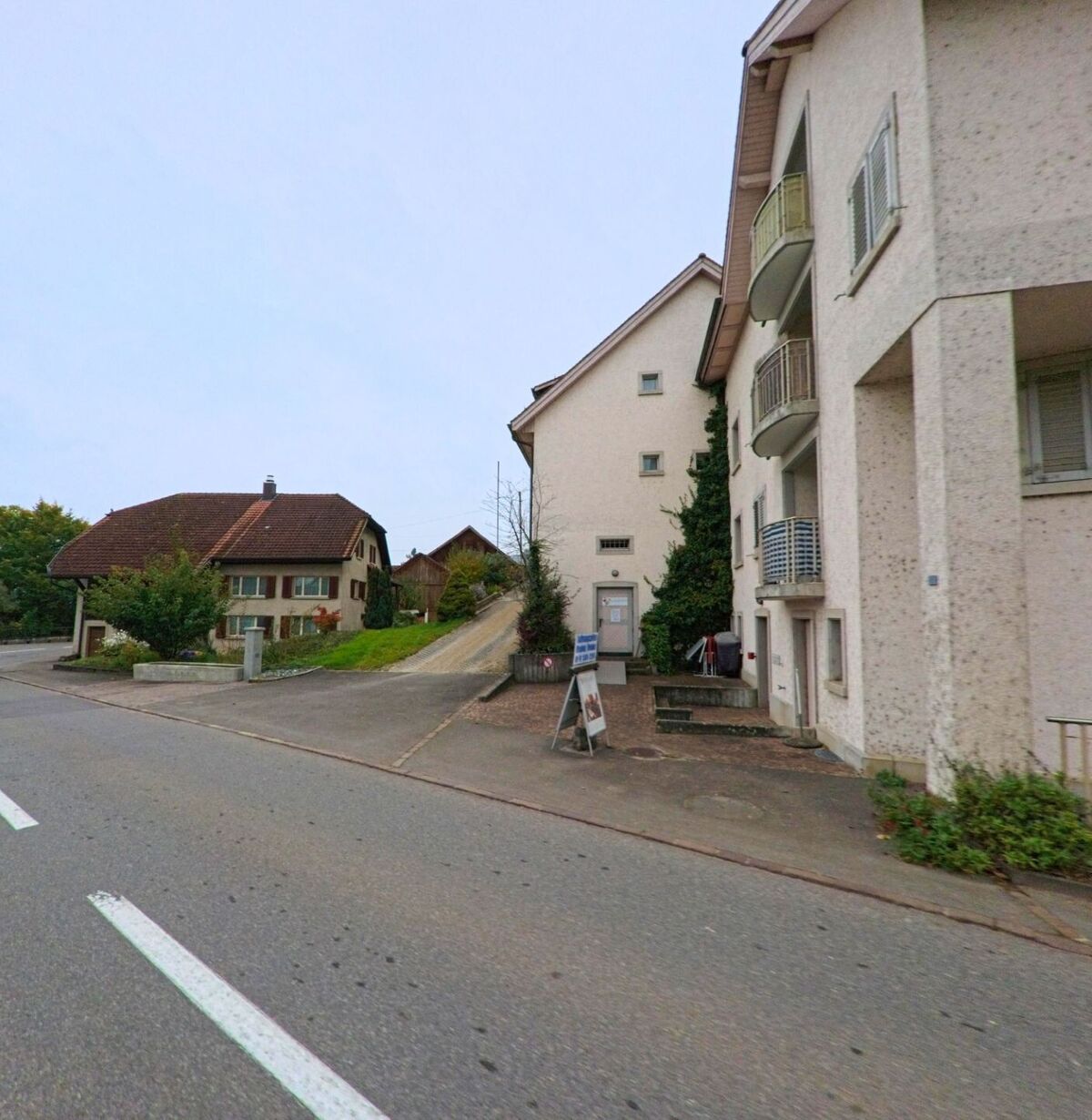 In Böttstein (5315, Kanton Aargau) stehen entlang der Hauptstrasse Wohnhäuser mit hellen Fassaden, während ein schmaler Weg zwischen den Gebäuden zu weiteren Häusern im Hintergrund führt.