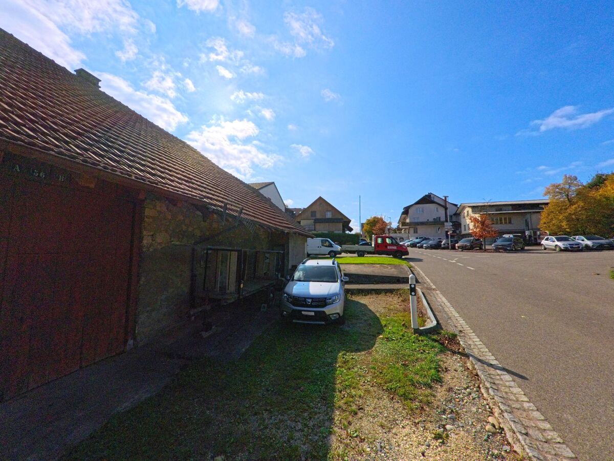 Ein älteres Steingebäude mit Ziegeldach steht in Bözberg (5225, Kanton Aargau) neben geparkten Autos, während sich im Hintergrund mehrere Wohnhäuser und Bäume zeigen.