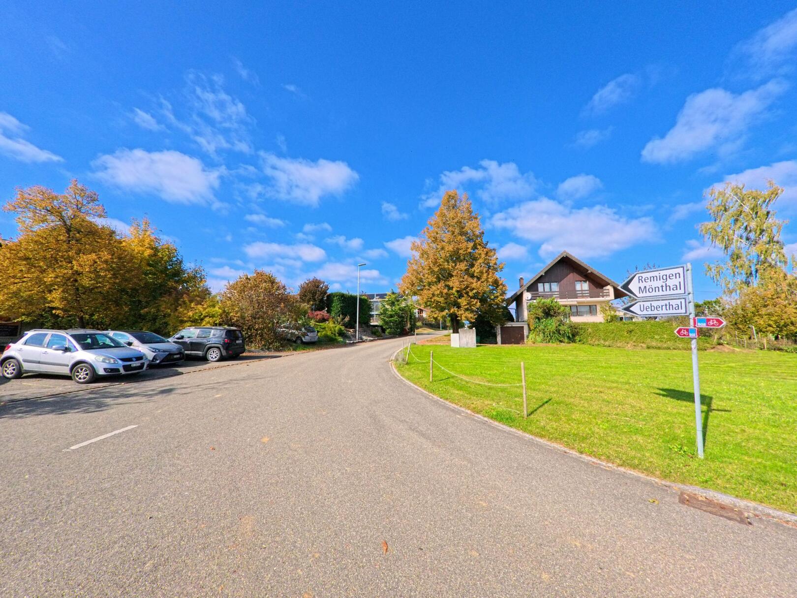 An einer ruhigen Strasse in Bözberg (5225, Kanton Aargau) weist ein Wegweiser Richtung Remigen, Mönthal und Ueberthal, während geparkte Autos und ein Einfamilienhaus das Dorfbild prägen.