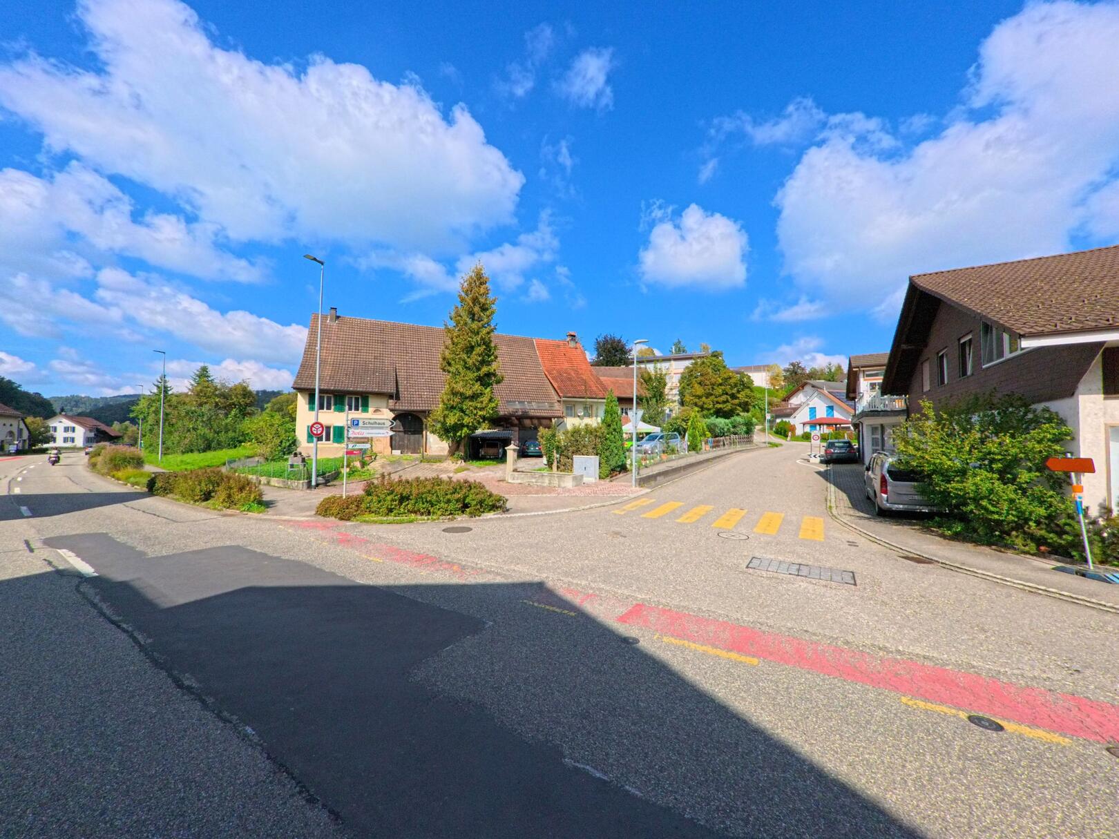 Eine Kreuzung mit Zebrastreifen und traditionellen Häusern prägt das Dorfbild von Böztal (5076, Kanton Aargau) an einem sonnigen Tag.