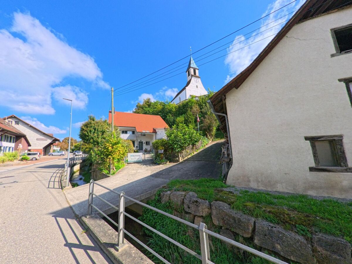 Ein schmaler Weg führt in Böztal (5076, Kanton Aargau) zwischen alten Häusern hinauf zur Kirche, die auf einer kleinen Anhöhe mit grünem Bewuchs steht.
