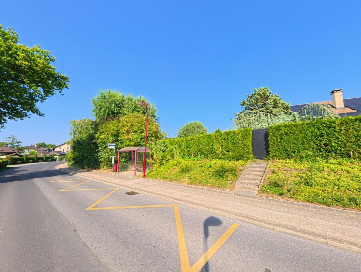 Eine Bushaltestelle mit Wartehaeuschen befindet sich in Bogis-Bossey (1279, Kanton Waadt) an einer Strasse, die von hohen Hecken und Gaerten umgeben ist.