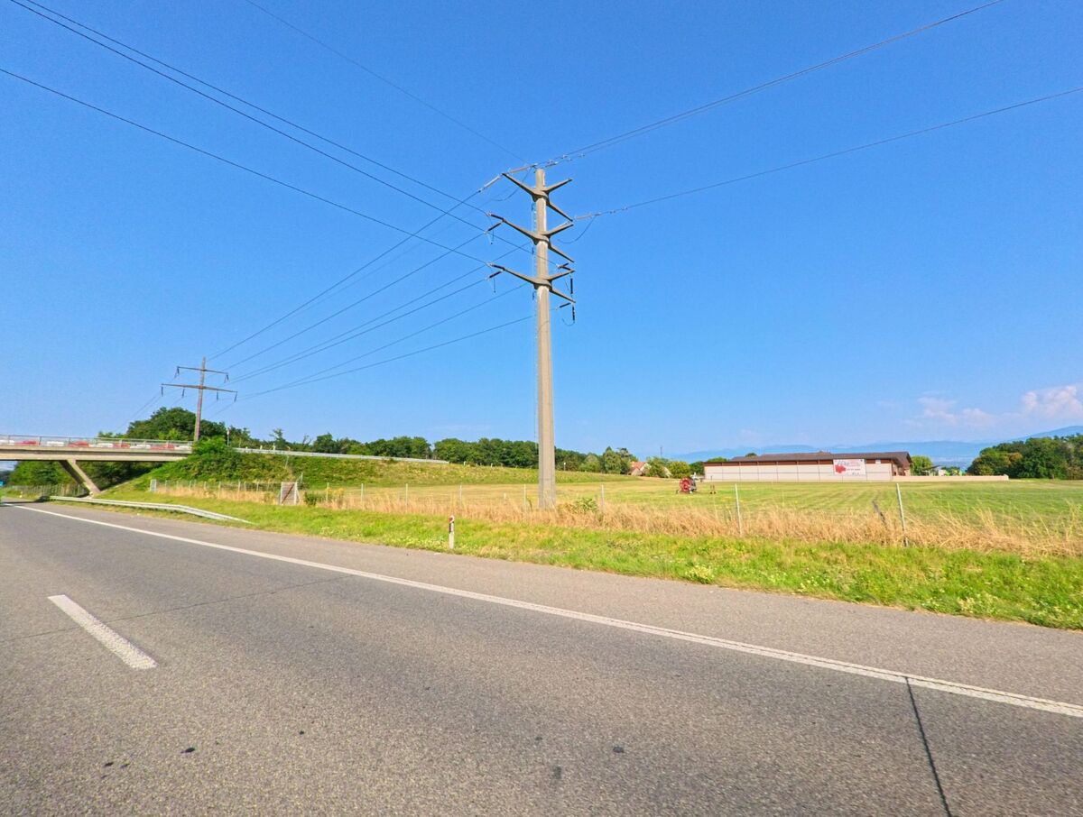 Neben der Hauptstrasse in Bogis-Bossey (1279, Kanton Waadt) verlaufen Stromleitungen ueber Felder, im Hintergrund ist eine Bruecke sichtbar.