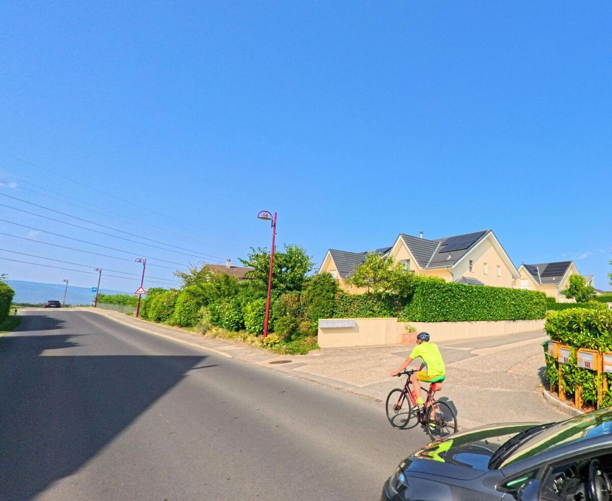 Ein Radfahrer faehrt durch ein Wohnquartier in Bogis-Bossey (1279, Kanton Waadt), waehrend im Vordergrund ein Auto mit geoeffnetem Fenster zu sehen ist.