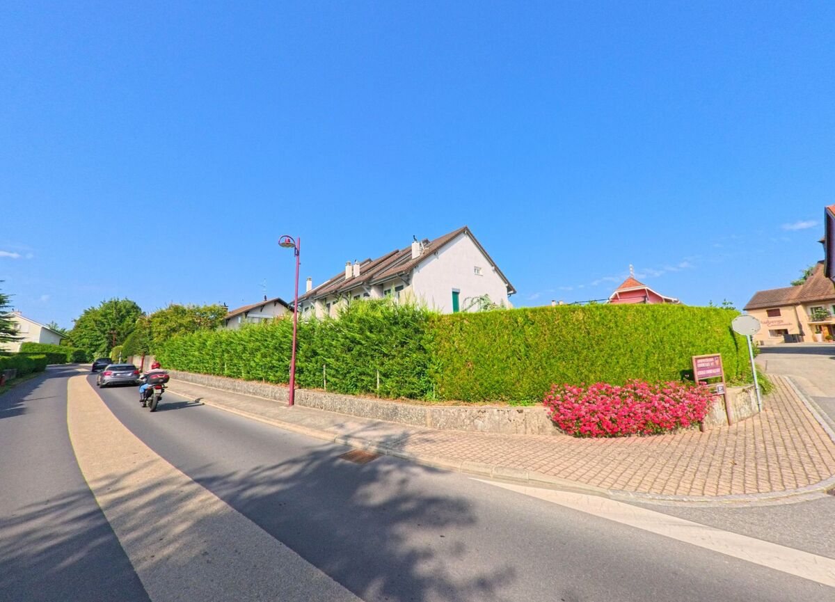 Eine Strasse in Bogis-Bossey (1279, Kanton Waadt) wird von Hecken und Wohnhaeusern gesaeumt, waehrend ein Motorradfahrer vorbeifaehrt.