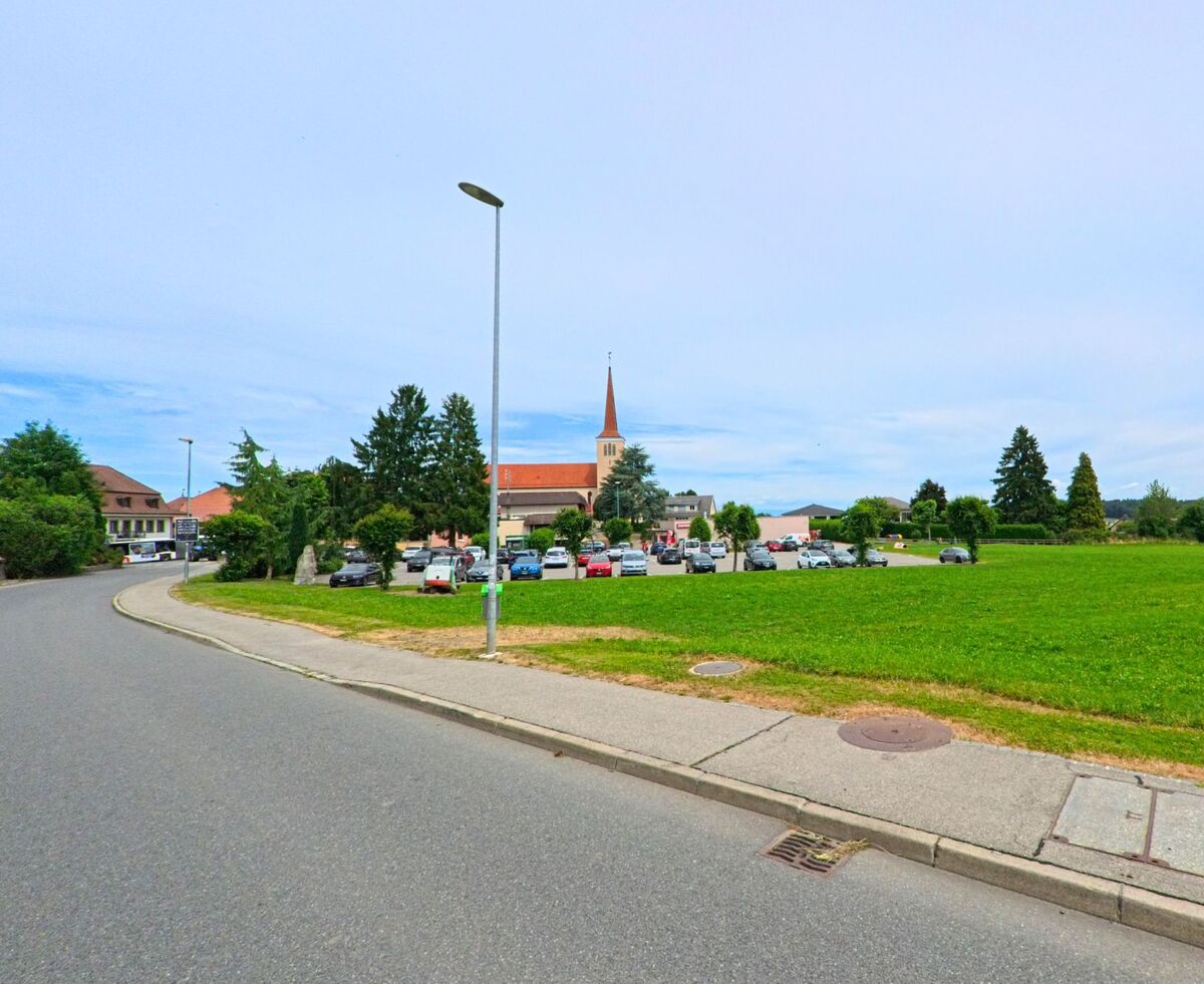 In Bois-d’Amont (1731, Kanton Freiburg) liegt ein Parkplatz mit zahlreichen Fahrzeugen vor einer Kirche mit spitzem Turm, angrenzend an eine grosse Wiese.