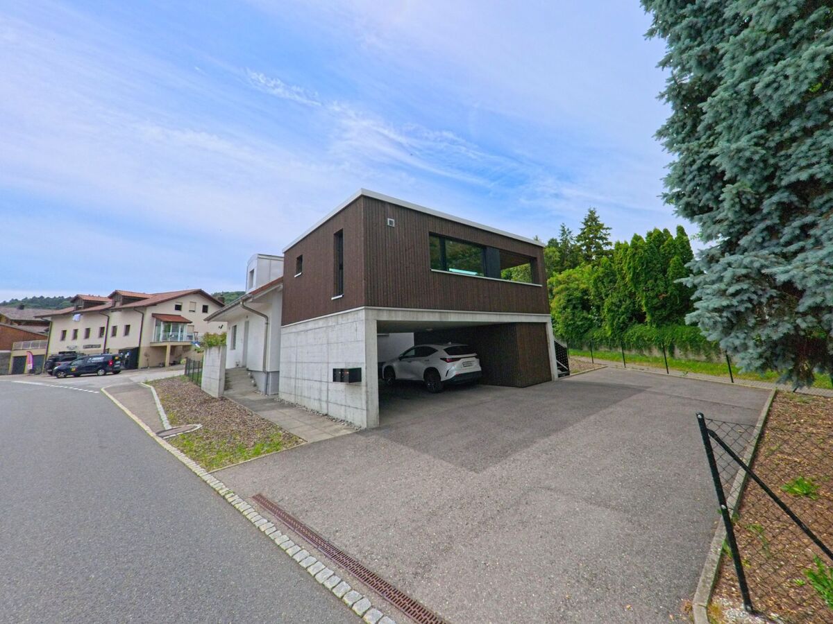 In Bois-d’Amont (1731, Kanton Freiburg) steht ein modernes Wohnhaus mit Carport an einer ruhigen Quartierstrasse, flankiert von hohen Bäumen und weiteren Einfamilienhäusern.