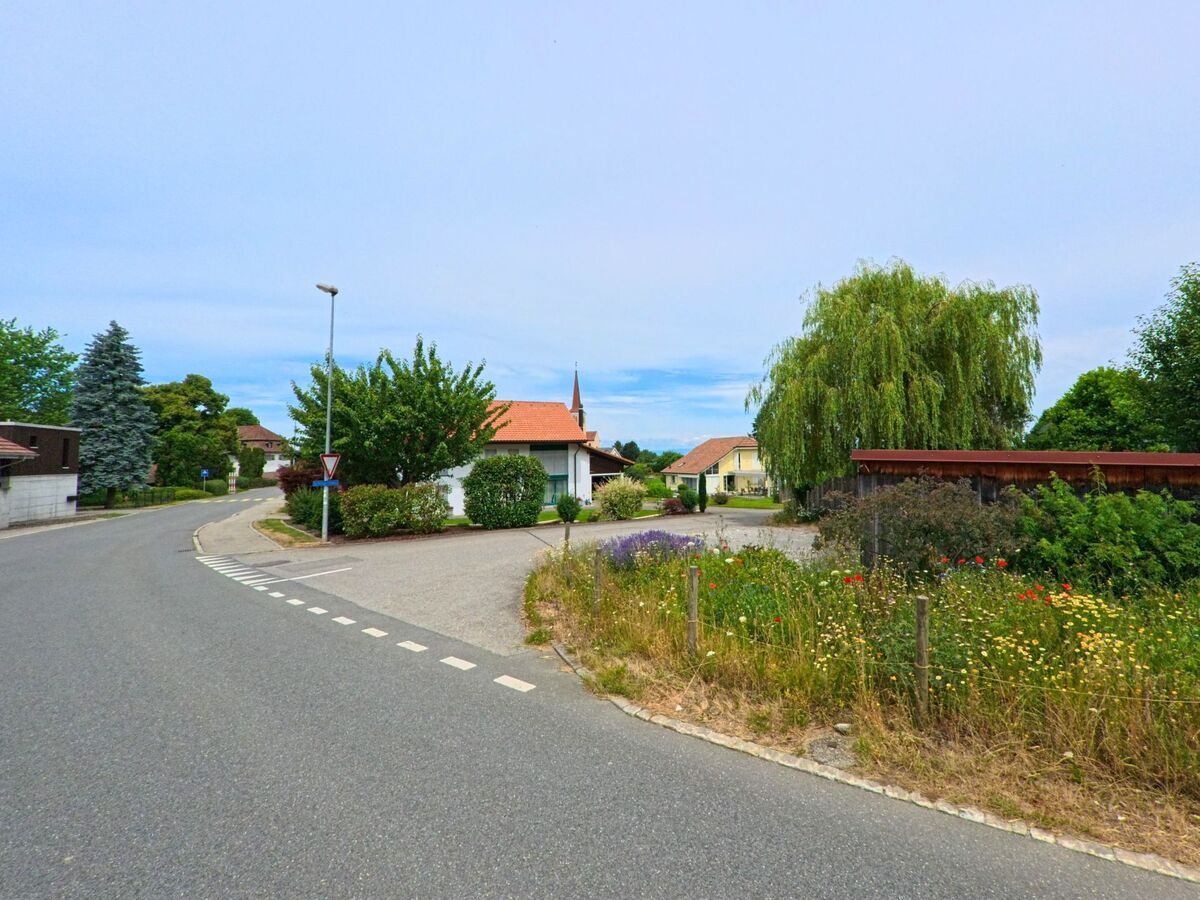 In Bois-d’Amont (1731, Kanton Freiburg) windet sich eine Wohnstrasse durch ein ruhiges Quartier mit Einfamilienhäusern, Wildblumenwiese und Blick auf einen Kirchturm.