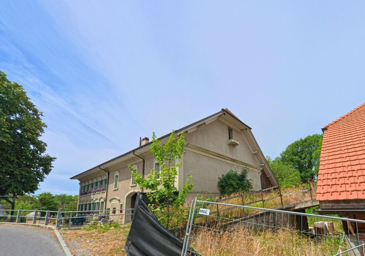 In Bois-d’Amont (1732, Kanton Freiburg) verläuft eine leicht ansteigende Strasse an einem historischen Gebäude und einem umzäunten Grundstück mit Neubau vorbei.