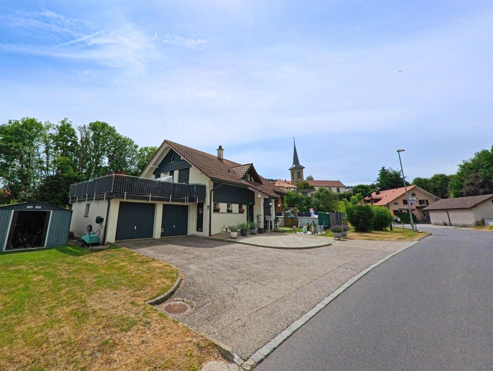 In Bois-d’Amont (1732, Kanton Freiburg) steht ein modernes Einfamilienhaus mit Garagenvorplatz und Garten, während im Hintergrund die Kirchturmspitze sichtbar ist.