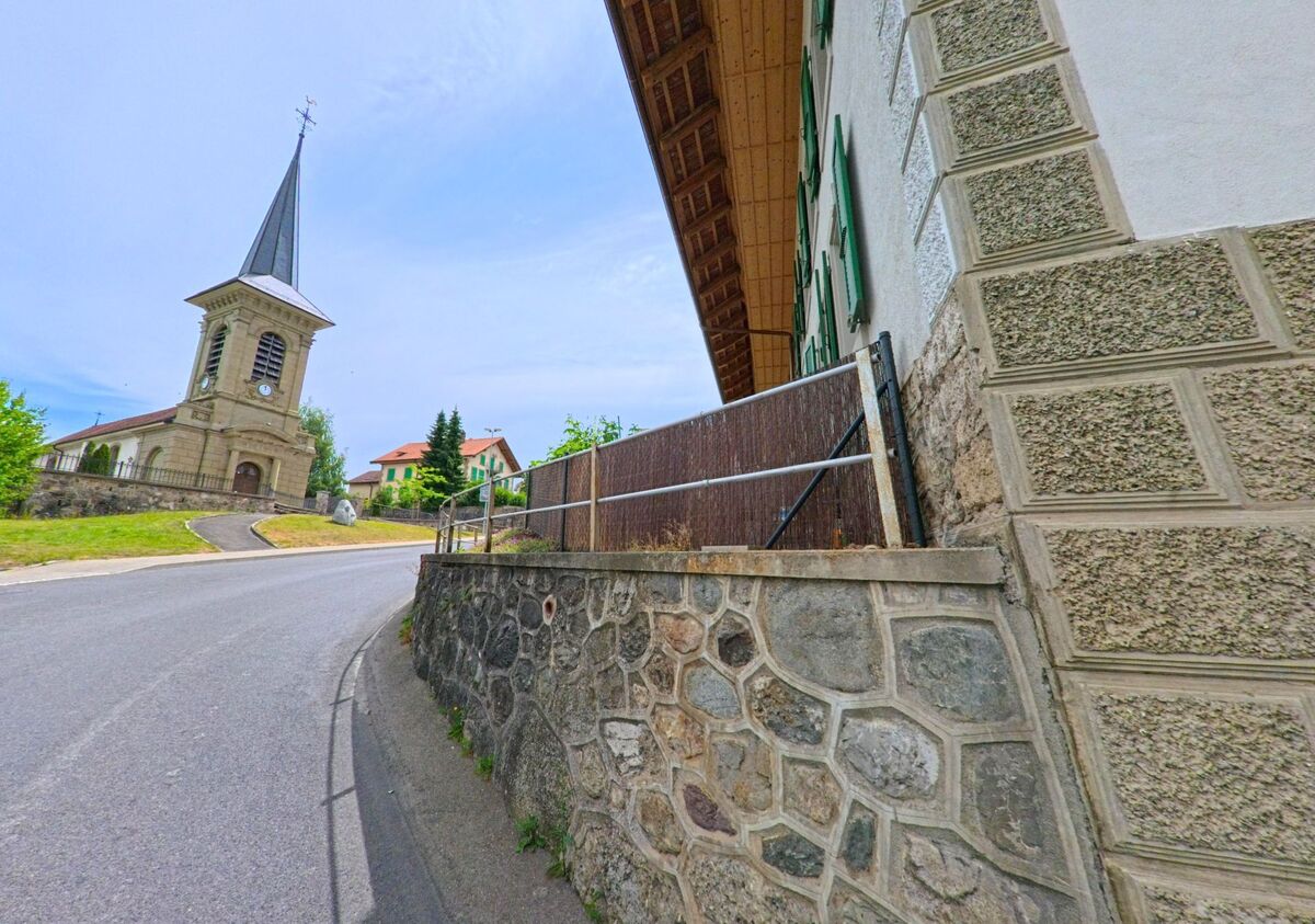 In Bois-d’Amont (1732, Kanton Freiburg) steigt eine kurvige Strasse zur Kirche mit markantem Turm auf, flankiert von Wohnhäusern und Natursteinmauern.