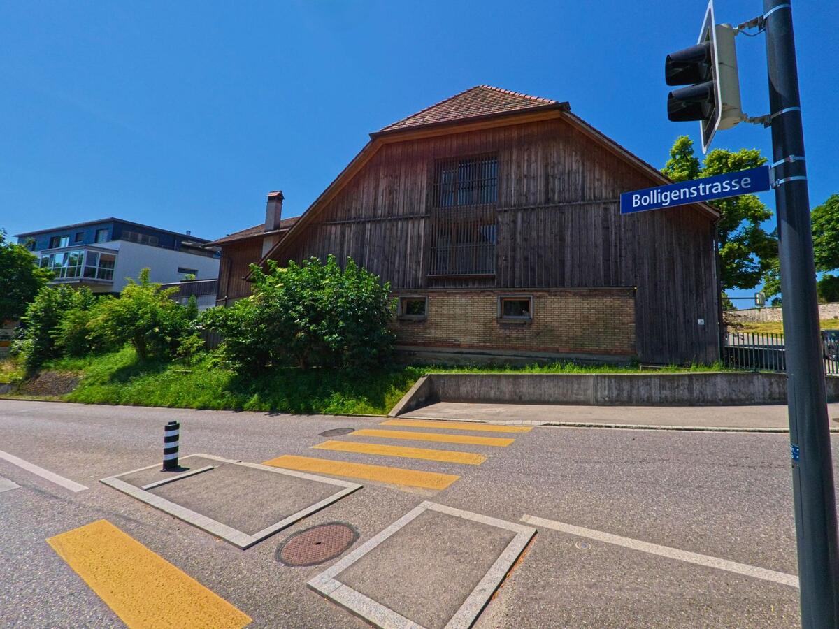Direkt an der Bolligenstrasse in Bolligen (PLZ 3065, Kanton Bern) steht ein traditionelles Holzhaus mit gemauertem Sockel neben einem Fussgaengerstreifen und einer Ampel.