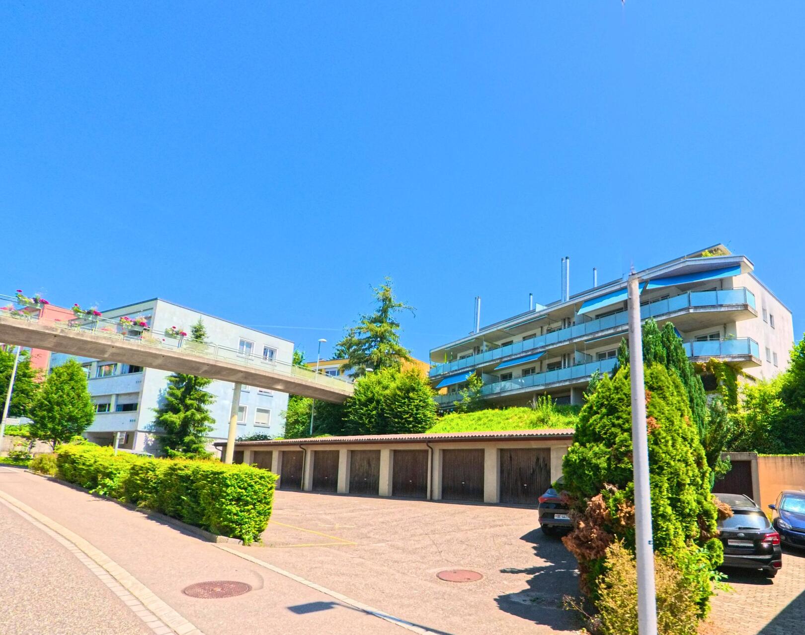 In Bolligen (3065, Kanton Bern) stehen moderne Mehrfamilienhaeuser oberhalb eines Parkplatzes mit Garagen und werden durch eine bepflanzte Fussgaengerbruecke verbunden.