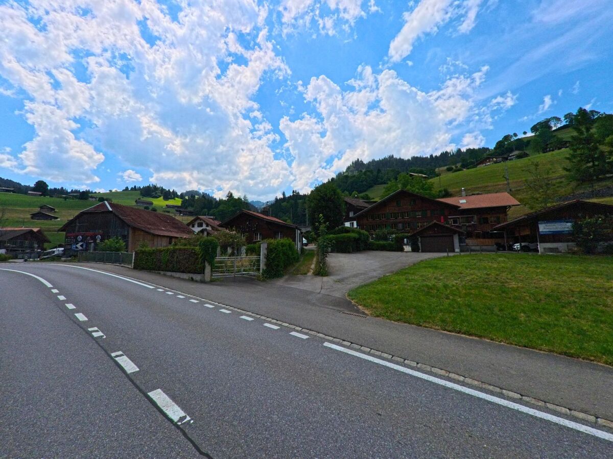 In Boltigen (PLZ 3766, Kanton Bern) führt eine sanft geschwungene Strasse an mehreren Holzhäusern vorbei, die am Fuss eines grünen Hangs mit verstreuten Einzelhöfen liegen.