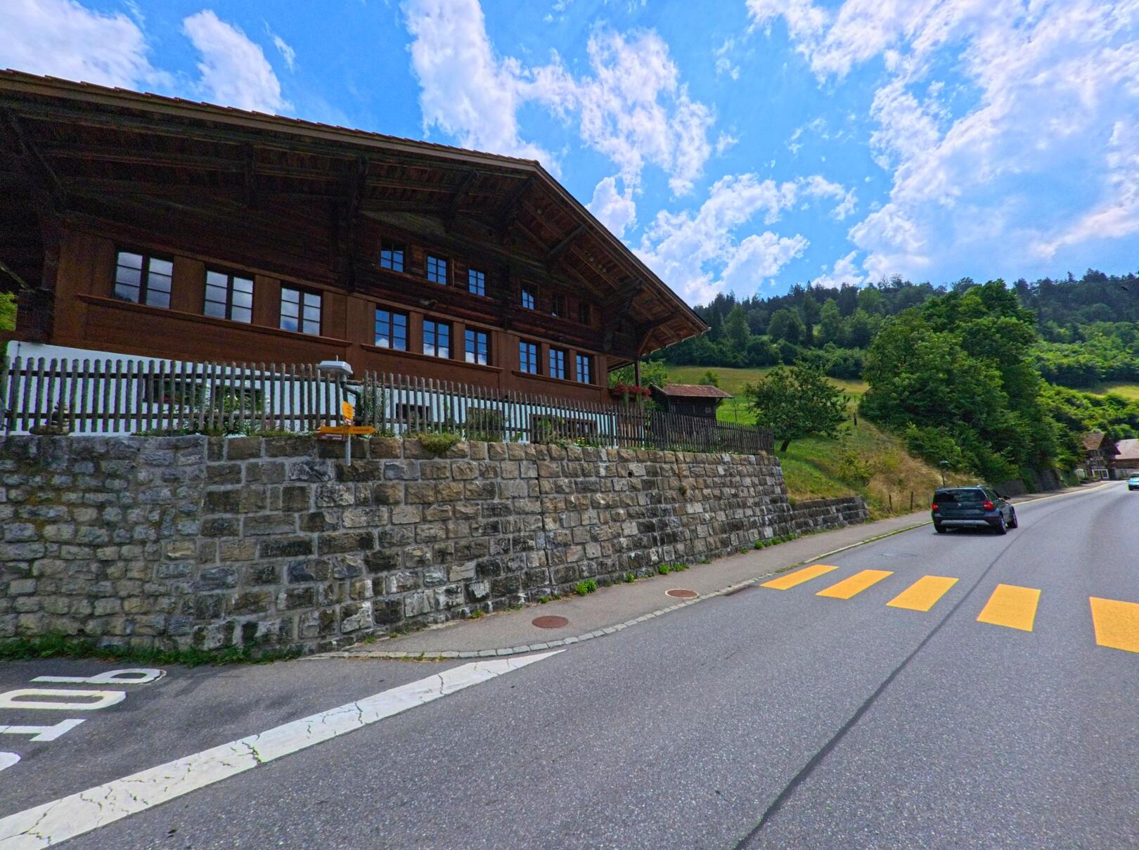 In Boltigen (PLZ 3766, Kanton Bern) verläuft eine Strasse an einem traditionellen Holzhaus mit steinernem Sockel und vorgelagerter Fussgängerstreifen entlang einer bewaldeten Hügelkulisse.