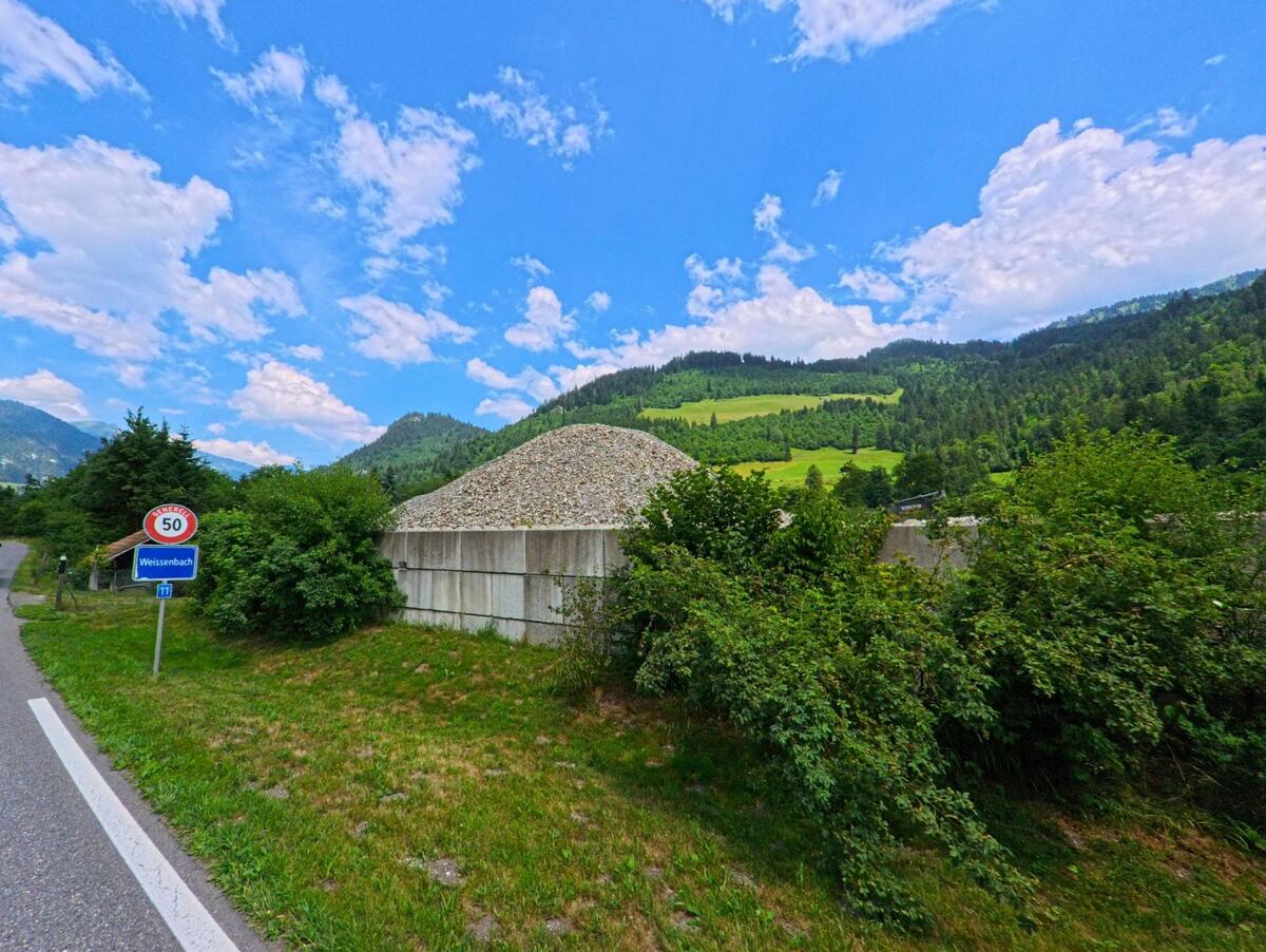 Am Ortseingang von Weissenbach bei Boltigen (PLZ 3766, Kanton Bern) liegt ein grosser Schotterhaufen hinter einer Betonmauer, umgeben von grünen Hügeln und Wald.