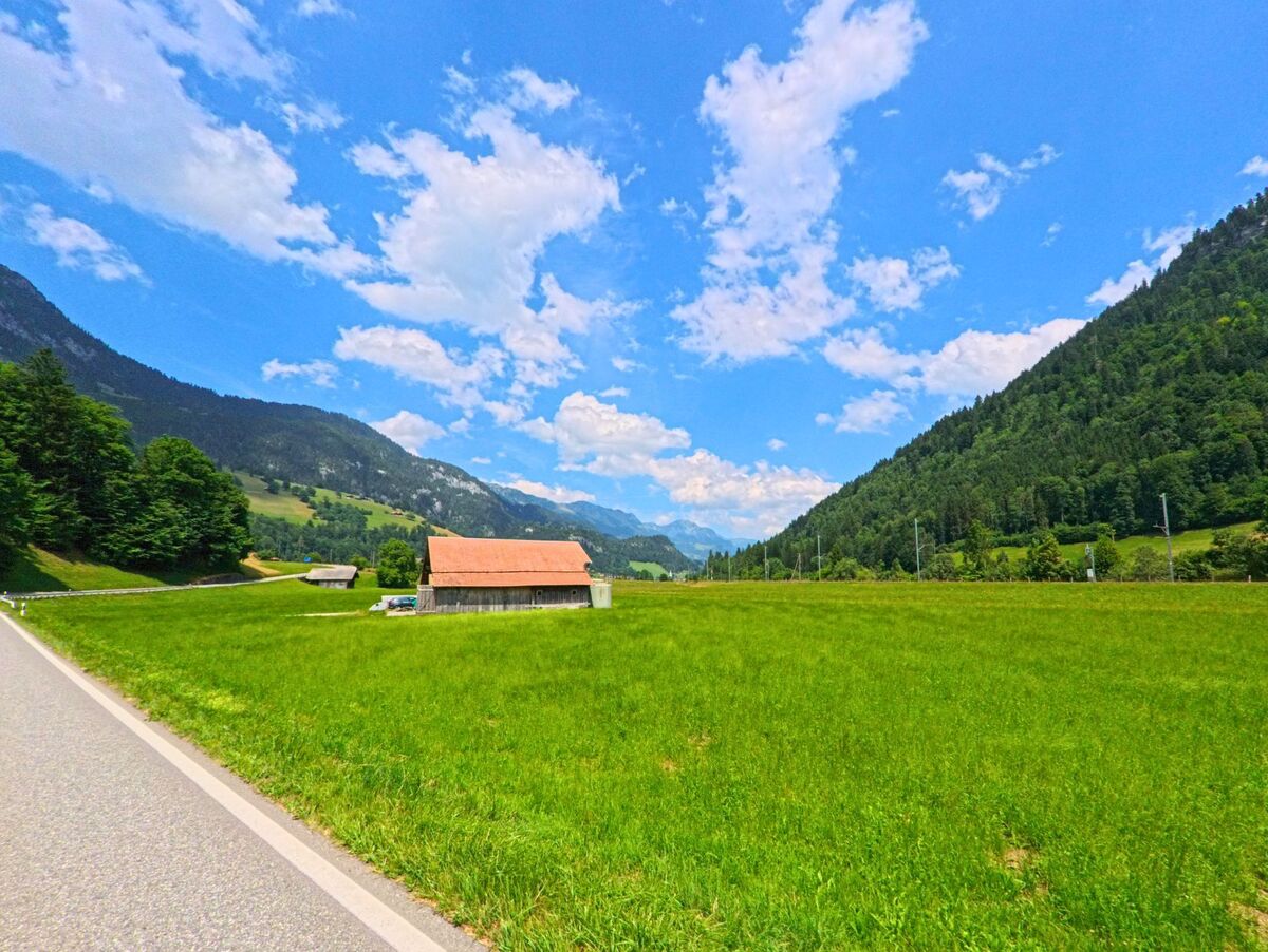 Inmitten weiter Wiesen von Boltigen (PLZ 3766, Kanton Bern) steht ein einfaches Wirtschaftsgebäude mit rotem Dach, eingerahmt von bewaldeten Berghängen und Fernblick in ein alpines Tal.