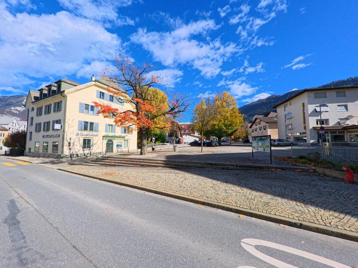 Am Rand eines kleinen Platzes in Bonaduz (7402, Kanton Graubünden) stehen Verwaltungs- und Wohngebäude neben herbstlich gefärbten Bäumen auf gepflastertem Untergrund.