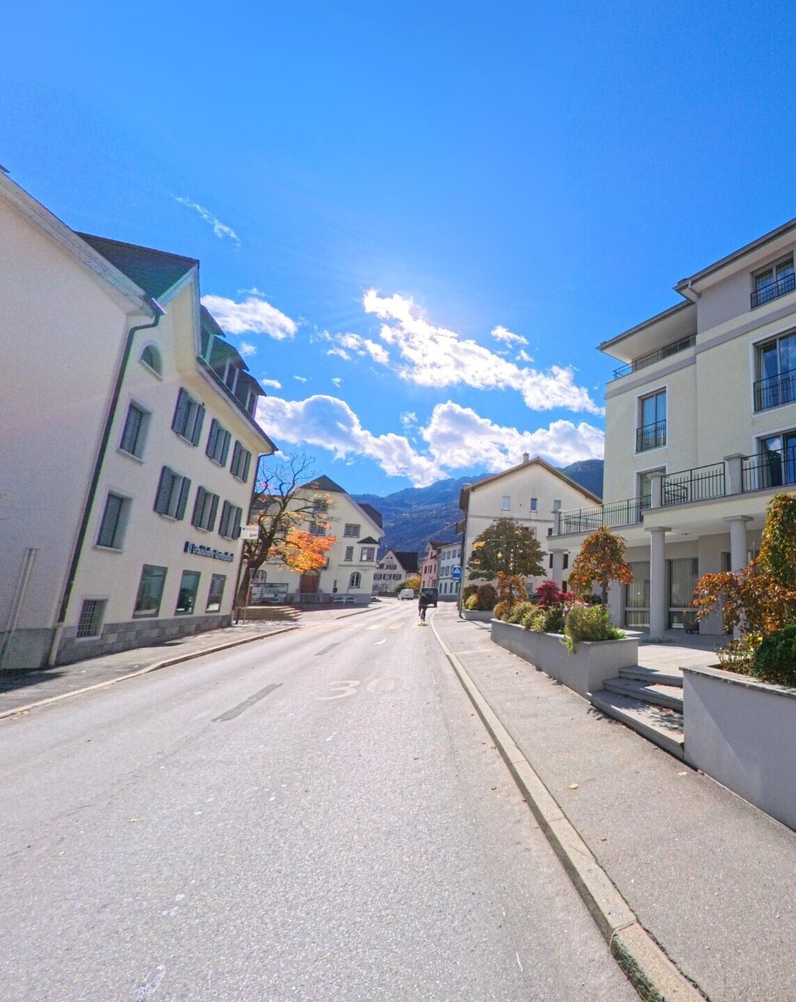 Eine gerade Dorfstrasse in Bonaduz (7402, Kanton Graubünden) führt zwischen Wohn- und Geschäftsgebäuden in Richtung der umliegenden Berge unter hellem Himmel.