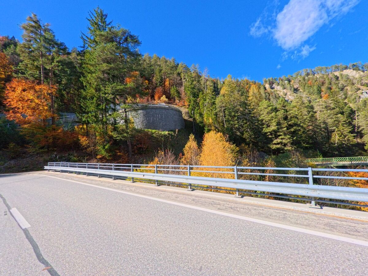 Entlang einer Strasse am bewaldeten Hang in Bonaduz (7402, Kanton Graubünden) verläuft eine Leitplanke vor herbstlich gefärbten Bäumen und einer steinernen Stützmauer.