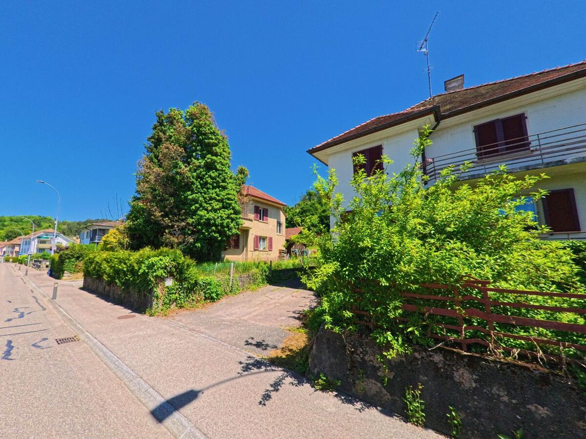 In Boncourt (PLZ 2926, Kanton Jura) stehen entlang einer sonnigen Strasse mehrere Wohnhäuser mit Vorgärten und dichter Bepflanzung.