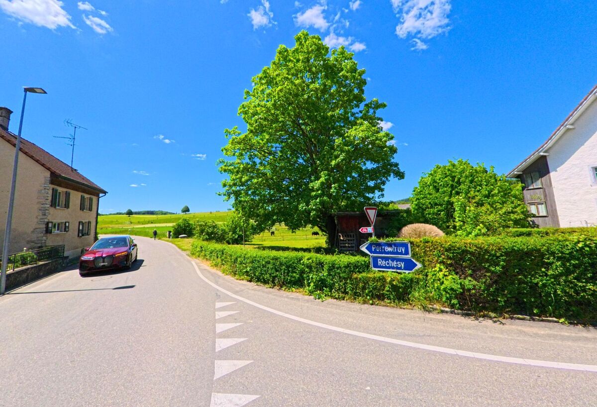 Eine Abzweigung in Bonfol (2935 im Kanton Jura) fuehrt mit Wegweisern Richtung Rechesy und Pfetterhouse, waehrend ein Auto auf der Hauptstrasse vorbeifährt.