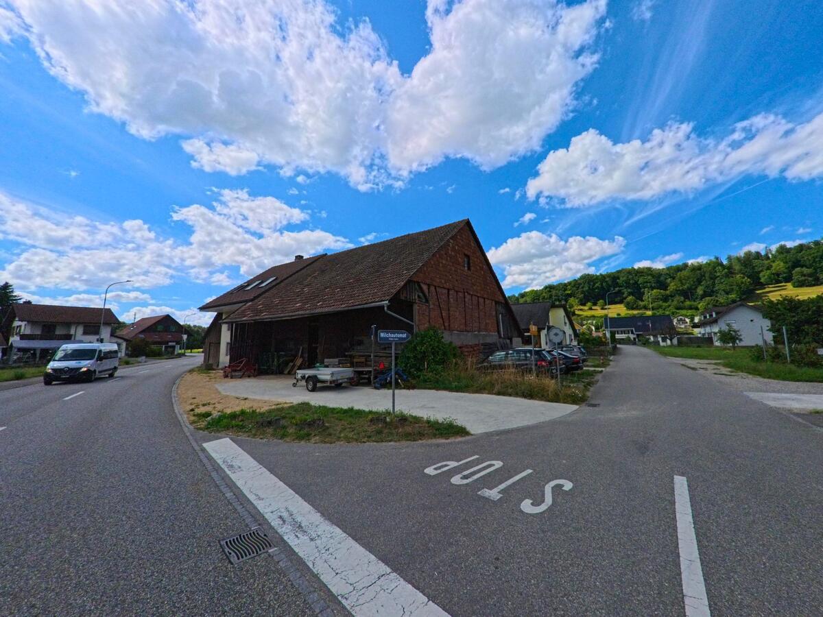 In Boningen (PLZ 4618, Kanton Solothurn) mündet die Milchstrasse an einem Bauernhaus in eine Hauptstrasse, während im Hintergrund grüne Hügel das Ortsbild prägen.