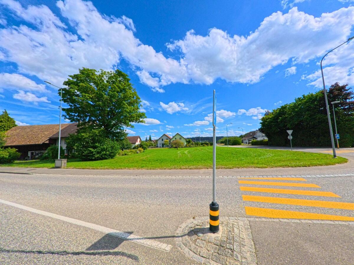 Ein gelb markierter Fussgaengerstreifen führt in Boningen (PLZ 4618, Kanton Solothurn) über eine Verkehrsinsel zwischen Wohnhäusern, Bäumen und einem kleinen Kreisel.