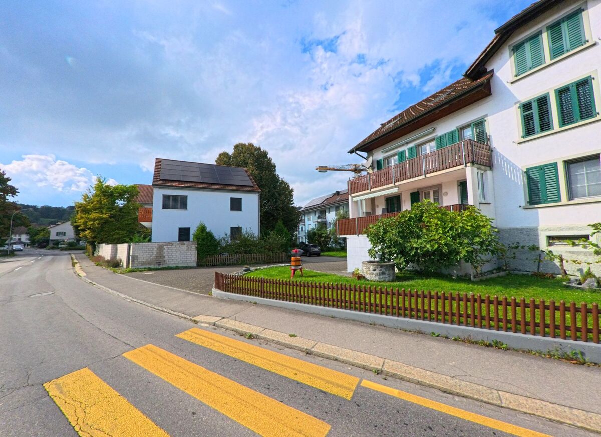 Entlang der Hauptstrasse in Boniswil (5706, Kanton Aargau) stehen gepflegte Wohnhäuser mit grünen Fensterläden, Vorgärten und einem Zebrastreifen im Vordergrund.